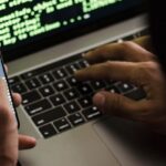 Malware - Unrecognizable hacker with smartphone typing on laptop at desk