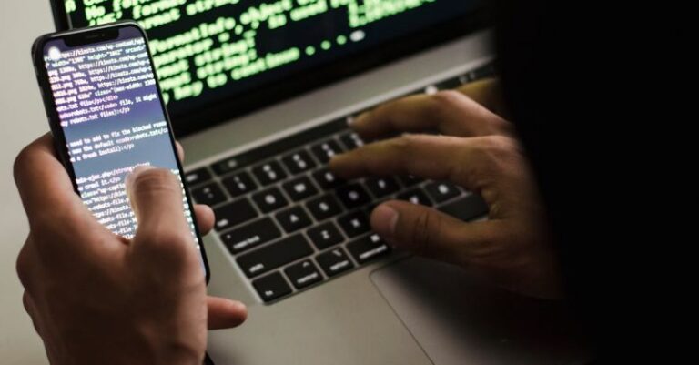 Malware - Unrecognizable hacker with smartphone typing on laptop at desk
