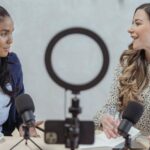 Hosting - Positive blogger asking questions to African American speaker and recording interview on cellphone at table with microphones